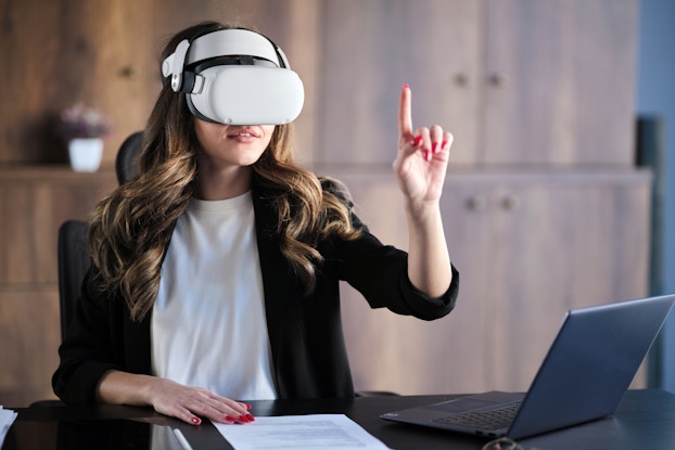 A woman wearing a VR headset sits at a desk and reaching up with one hand, a finger extended to poke or tap at something that only she can see. The table she sits at also holds an open laptop and a piece of paper covered with printed text.