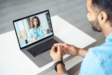  An over-the-shoulder shot of a video call taking place between a man and his doctor. The doctor can be see on the screen of an open laptop; she has dark curly hair and is wearing a lap coat with a stethoscope draped around her neck. The man is seen from behind, partially off-screen and out-of-focus; he has dark hair and a dark beard and he is wearing a chambray shirt. 