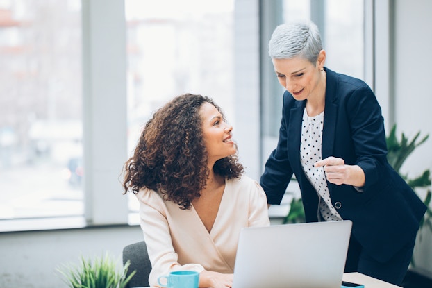  An older, short-haired woman leans over a younger woman sitting at a laptop. The younger woman looks up at the older woman, who appears to be explaining something.