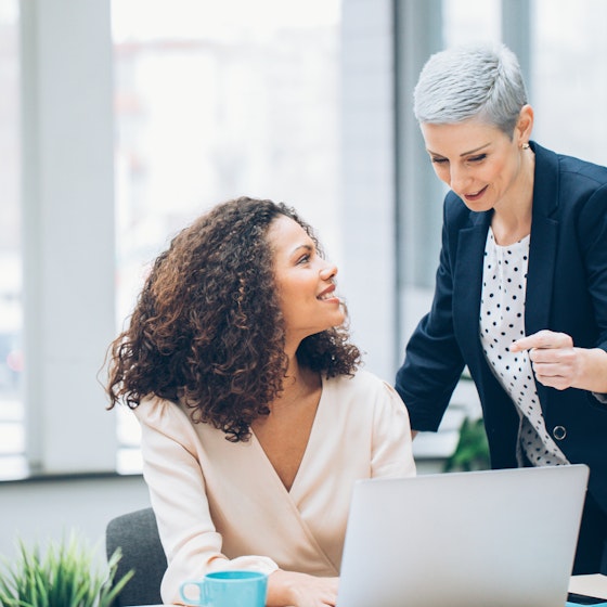 An older, short-haired woman leans over a younger woman sitting at a laptop. The younger woman looks up at the older woman, who appears to be explaining something.