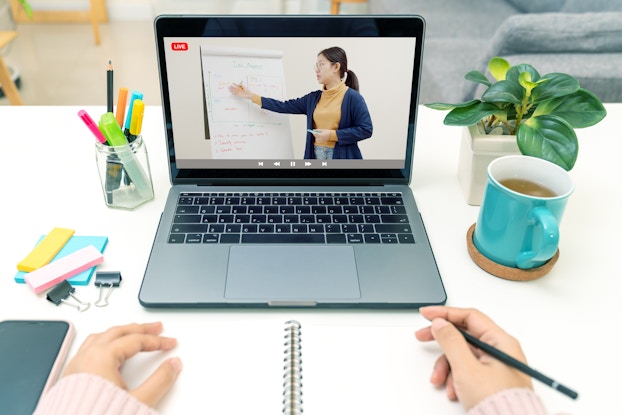  A POV shot of someone watching a class on a laptop and taking notes in a notebook. The laptop screen shows a woman gesturing to an easel displaying a large pad of paper covered with notes.