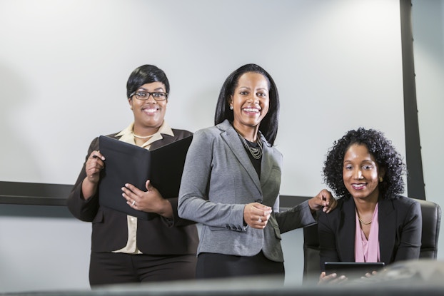 A trio of Black women wearing business attire faces the camera. The women are smiling. The woman standing on the left is holding an open binder and the one sitting on the right is holding an electronic tablet.