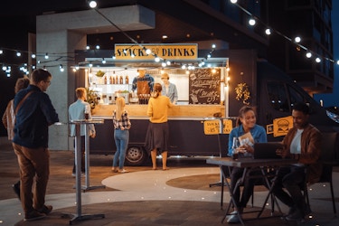  A food truck is parked on a concrete plaza under strings of lights. Three patrons line up before the truck, which advertises SNACKS AND DRINKS. Other customers stand and sit at metal tables set up around the plaza. 