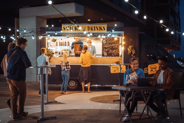  A food truck is parked on a concrete plaza under strings of lights. Three patrons line up before the truck, which advertises SNACKS AND DRINKS. Other customers stand and sit at metal tables set up around the plaza.