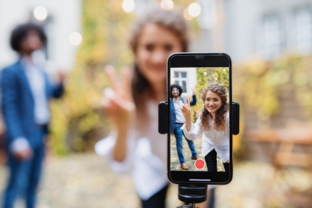  In the foreground and slightly right of center, a smartphone in a tripod holder shows a woman and, a few feet behind her, a man. Both of them are standing outside in front of a wall draped in ivy, making peace signs at the camera. In the background out of focus, the woman and man pose for the camera.