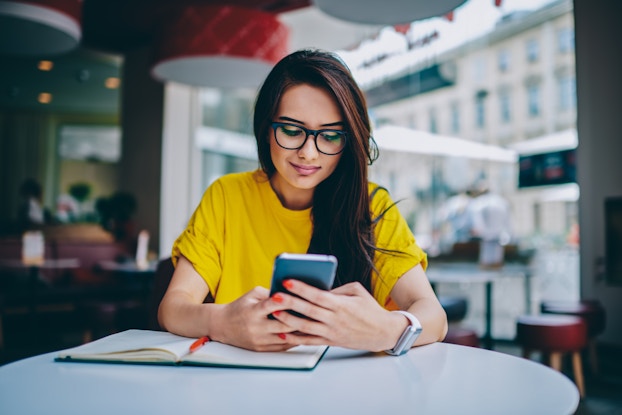 A young woman wearing thick eyeglasses and a yellow T-shirts sits at a round table in a restaurant or cafe and looks at her smartphone with a slight smile on her face.