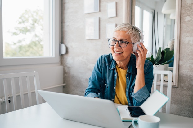  An older women with short white-blond hair sits at a table in front of an open laptop, an open book, a mug and a smartphone. She wears a set of headphones and has one hand up to hold them in place.