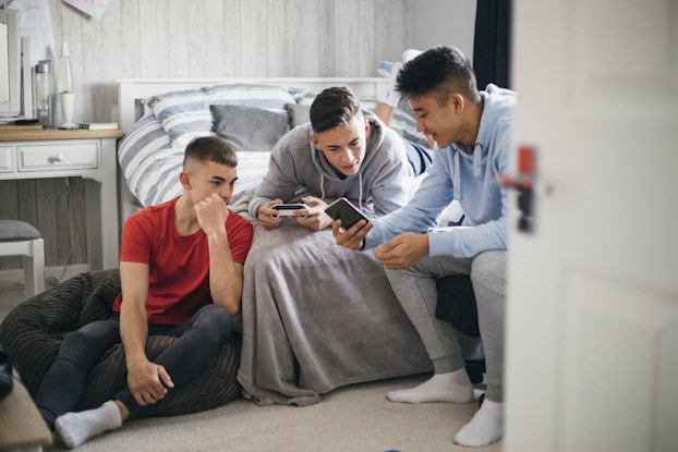  A trio of teenage boys sit on and around a bed in a bedroom with a white-and-gray color scheme. The boy on the right, wearing a light blue hoodie, shows something on his smartphone to the other two boys. The boy in the middle is wearing a gray hoodie and holding a handheld game console. The boy on the left is sitting on the floor, wearing a red T-shirt and holding one hand to his chin.