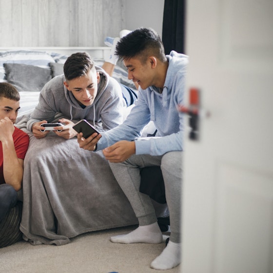  A trio of teenage boys sit on and around a bed in a bedroom with a white-and-gray color scheme. The boy on the right, wearing a light blue hoodie, shows something on his smartphone to the other two boys. The boy in the middle is wearing a gray hoodie and holding a handheld game console. The boy on the left is sitting on the floor, wearing a red T-shirt and holding one hand to his chin. 