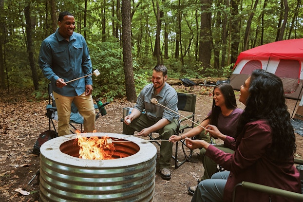  A group of friends roasts marshmallows around a metal firepit. They're sitting in foldable chairs in a forest clearing. A large tent is set up in the background.