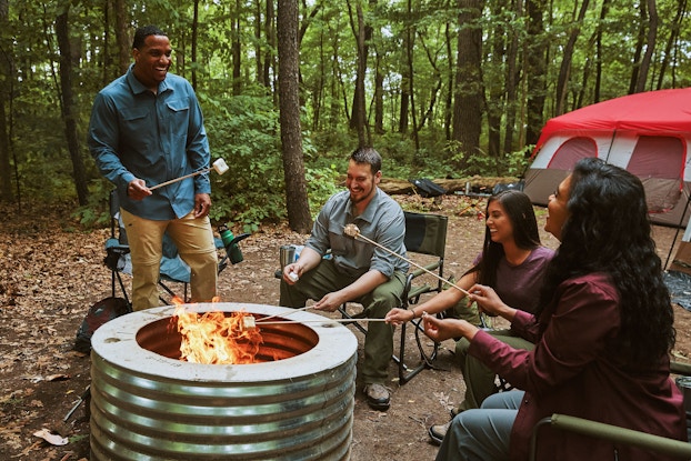 A group of friends roasts marshmallows around a metal firepit. They're sitting in foldable chairs in a forest clearing. A large tent is set up in the background.