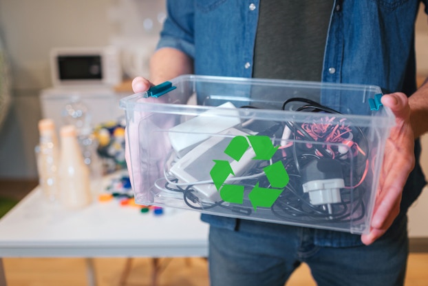  A close-up shot of a person seen from the shoulders-down holding a transparent box filled with wires, chargers, cell phone boxes and wired headphones. The box has a green recycling symbol on one side.