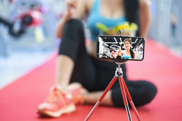 A smartphone is positioned horizontally on a tripod. On the screen of the phone is an image of a woman holding up a forkful of salad. The woman has her dark hair pulled back in a ponytail and is wearing a light blue workout crop top. The woman appears to be in a gym; behind her are several workout machines. In the background beyond the phone and tripod, out of focus, is the same woman on the screen, sitting on the floor of a gym with workout machines in the background.