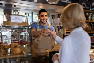 A young male bakery shop owner smiles as he hands a bagged item to a customer.
