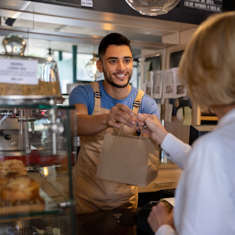 A young male bakery shop owner smiles as he hands a bagged item to a customer.