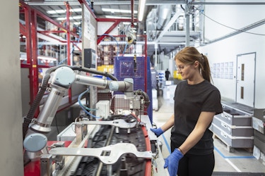 A young woman in a high-tech automotive parts factory watches a robotic arm drill holes in a car part. The woman has dark blonde hair pulled back into a ponytail, and she wears a black T-shirt, black pants, and blue rubber gloves. The factory room is cavernous with white walls, bright overhead lights, and red metal beams partitioning the floor into different areas. 