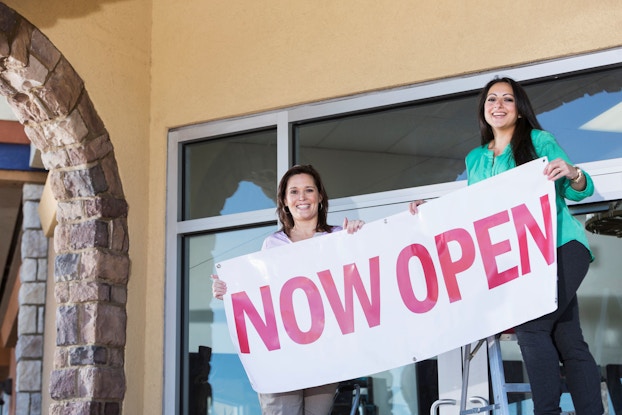  Two women entrepreneurs hang a Now Open sign in front of their storefront.