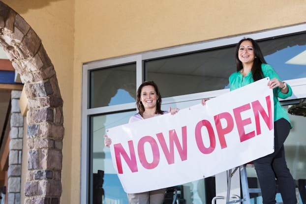 Two women entrepreneurs hang a Now Open sign in front of their storefront.