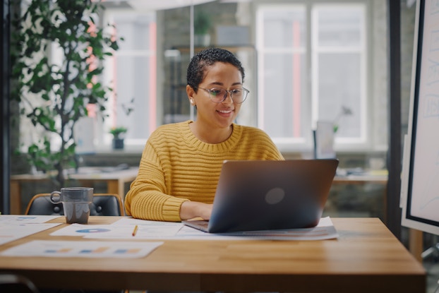 A young grinning woman sits at a wood-topped table and works on a laptop. The woman wears a yellow sweater and round glasses. Several papers are scattered on the table and a whiteboard stands next to the table. Behind the woman is a tall potted plant and another long table, beyond which is a wall of windows.