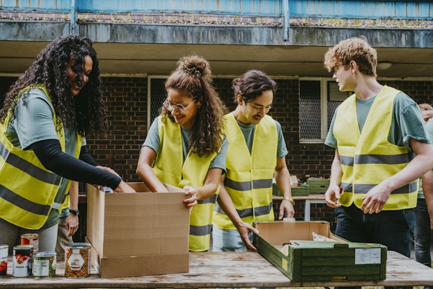 Four people wearing green T-shirts and high-visibility yellow vests stand at a table outside a building, packing cardboard boxes. The two people on the left, both women with long curly hair, are packing a box with cans of food. The two people on the right, both men, are speaking to each other while the shorter man on the left looks down at a long, flat box.