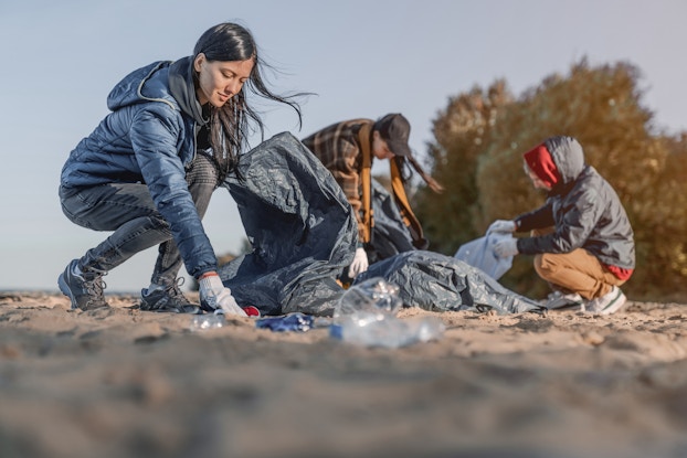 Three people dressed for cold weather pick up trash on a beach. Each person wears light gray gloves. The person closest to the camera is a long-haired woman wearing a blue jacket; she is crouched down, picking up a crushed red cup with one hand and holding a large black garbage bag in the other hand. The two people further in the background are out of focus. The one on the left is wearing a plaid jacket and brown scarf and is bending over to pick up a garbage bag. The one of the right is wearing a black-and-red windbreaker and is crouched on the ground, opening up a new garbage bag.