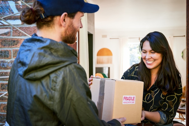  A deliveryman hands a package to a smiling woman standing in an open doorway. The package is a square cardboard box with a sticker reading "FRAGILE" on the side. The man has a reddish beard and reddish hair pulled into a bun under a navy blue cap. He wears a black windbreaker. The woman has long dark hair and wears a dark brown blouse covered with a gold squiggly design.