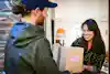 A deliveryman hands a package to a smiling woman standing in an open doorway. The package is a square cardboard box with a sticker reading "FRAGILE" on the side. The man has a reddish beard and reddish hair pulled into a bun under a navy blue cap. He wears a black windbreaker. The woman has long dark hair and wears a dark brown blouse covered with a gold squiggly design.