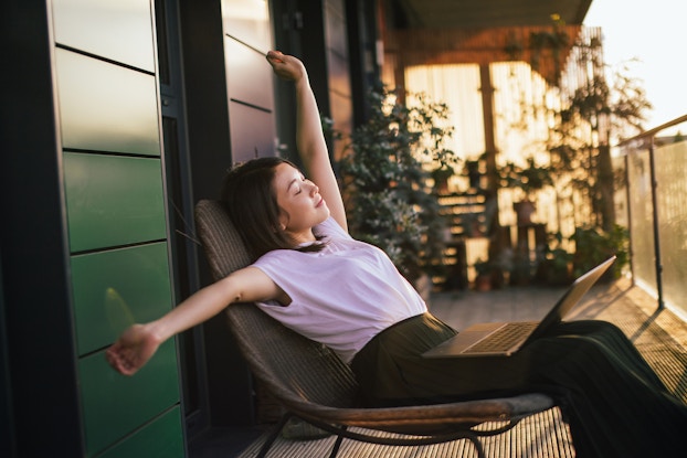 Woman stretching in a chair out on a deck with a laptop on her lap.