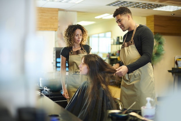 Two hair stylists stand over a client in a styling chair. The stylist on the right is a tall man in a long-sleeved black shirt and khaki apron; he stands behind the client and holds the ends of her long brown hair. The stylist on the left, a curly-haired woman also wearing a khaki apron, observes.