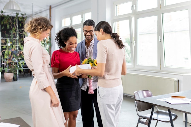 A group of office employees welcome a new colleague with flowers and a present.