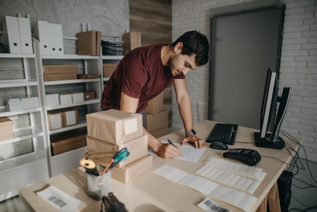 A man stands over a table in a storage room. He writes something on a piece of paper. On the table around him are packages, a desktop monitor and keyboard, a credit card reader, and various receipts.