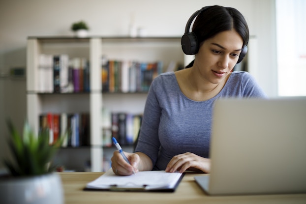 A woman wearing headphones watches something on a laptop. She takes notes on a clipboard next to her on the desk.