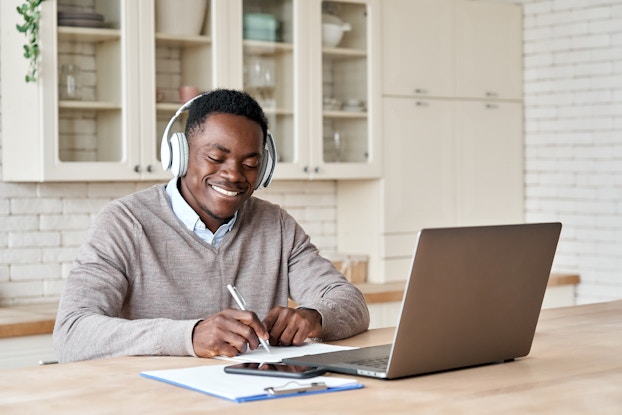  A smiling man sits at a wood-topped counter in front of an open laptop. He wears white headphones and writes something on a piece of paper. In the background are glass-fronted cabinets filled with dishes, jars and other kitchenware.