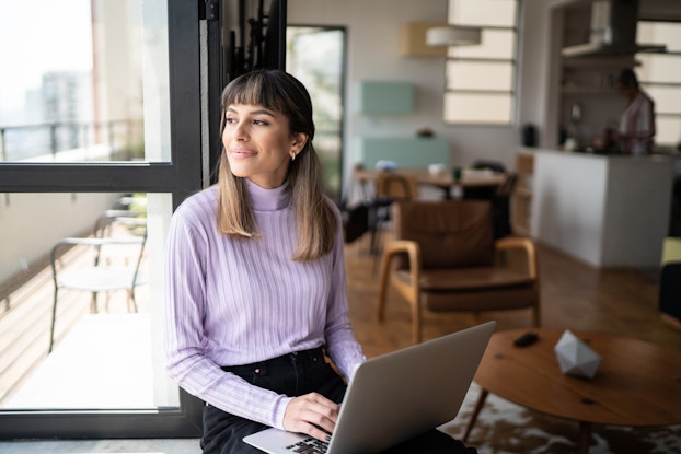A woman sits on a windowsill and looks offscreen with a hopeful smile on her face. She holds a laptop in her lap and has her hands on the keybaord. Behind her, the window looks out onto a long balcony. The woman has brown hair and wears a lilac sweater and black pants.