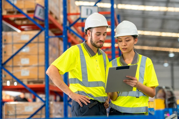 Two warehouse workers wearing bright yellow high-visibility polo shirts and white hard hats look at a clipboard. In the background are rows of floor-to-ceiling metal shelving units holding large pallets of boxes.