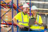 Two warehouse workers wearing bright yellow high-visibility polo shirts and white hard hats look at a clipboard. In the background are rows of floor-to-ceiling metal shelving units holding large pallets of boxes.