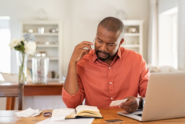  A bearded man in an orange button-up shirt sits at an open laptop and looks down at an open notebook with concern. With one hand, he holds the tip of a pen to the side of his head, as if scratching his head in confusion. In his other hand, he holds a small index card. In the background is a white minimalist room with two white bookcases and a table holding a vase of white flowers.