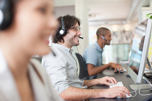  A man wearing a headset with a mouthpiece is looking at a large computer monitor and smiling. His hands are positioned on a keyboard attached to the monitor. In the foreground and background, out of focus, are two other people wearing similar headsets and working on simliar computers.