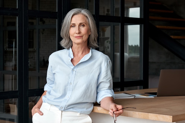  A older woman with shoulder-length gray hair leans casually against a wooden-topped counter. In the background is a dimly lit room behind glass walls and a staircase leading upstairs.