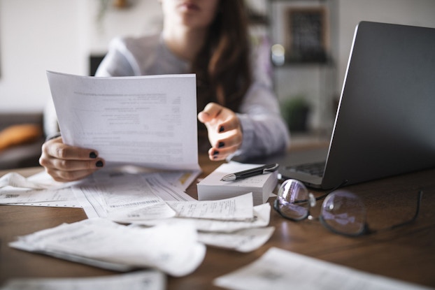 Close-up of a woman who is reviewing and revising her budget. She is holding up papers and has receipts spread out in front of her in addition to a notepad, a pen, and a laptop.