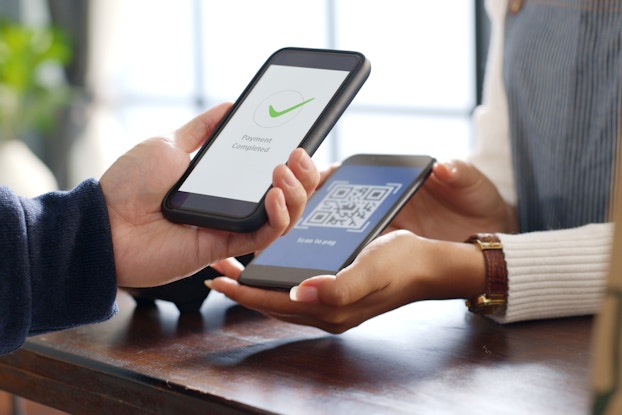 Two sets of hands hold smartphones near each other. The smartphone on the left, held slightly higher, shows a white screen with a green checkmark and the caption "Payment Completed." The smartphone on the right, held by a hand with French manicured nails, shows a QR code against a blue background.