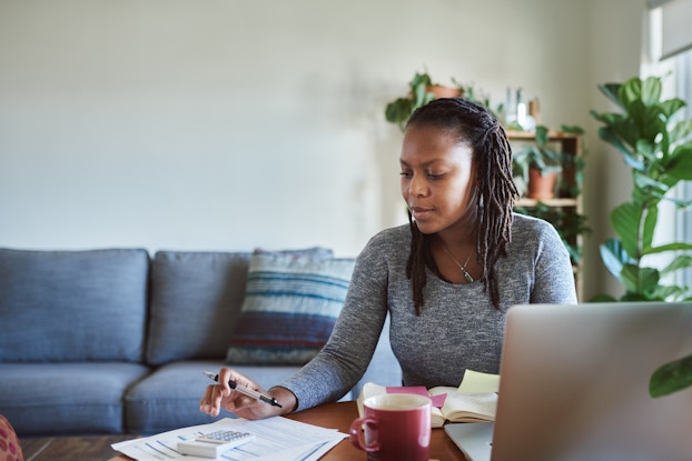 A woman sits at a coffee table in a living room. In front of her on the table are an open laptop, a coffee mug on a coaster and a stack of papers topped with a calculator. The woman looks at the papers and uses her right hand to both hold a pen and type something on the calculator.