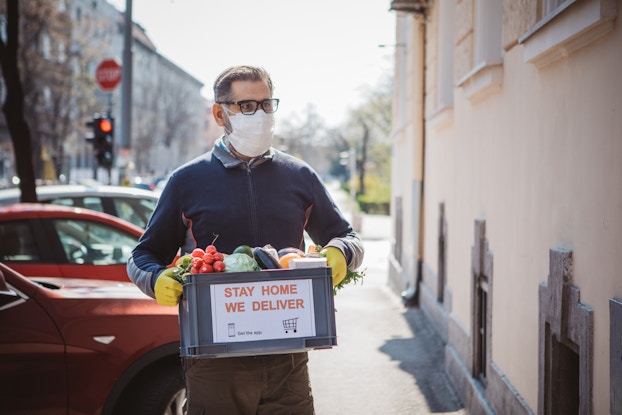 A man wearing a face mask and rubber gloves carries a box of vegetables away from a line of parked cars. A sign on the box reads "STAY HOME. WE DELIVER."