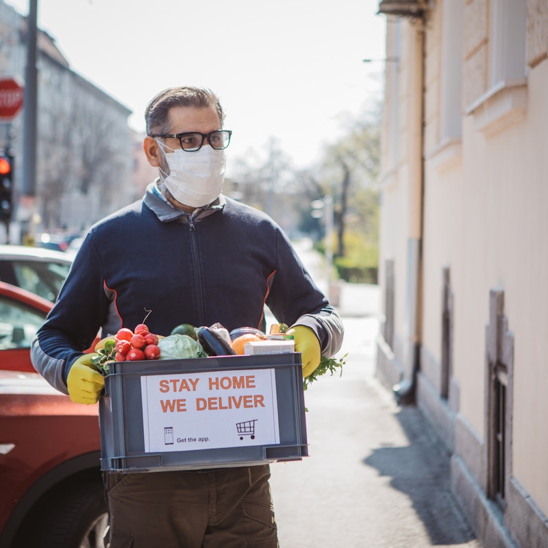 A man wearing a face mask and rubber gloves carries a box of vegetables away from a line of parked cars. A sign on the box reads "STAY HOME. WE DELIVER."