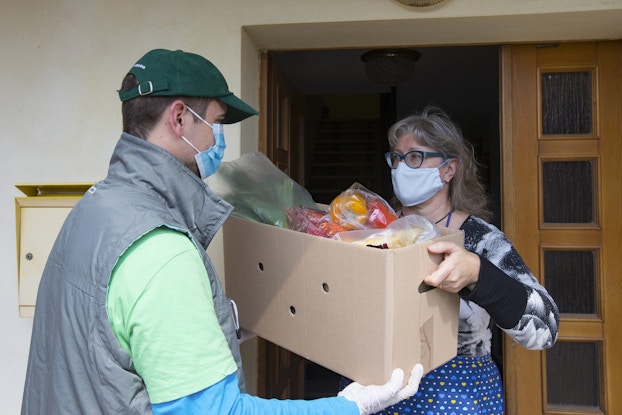  A man in a face mask and ball cap hands a box of groceries to a woman who also wears a face mask.