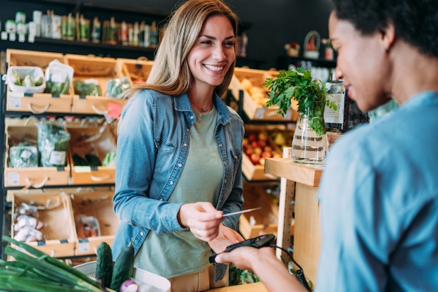 A woman holds out her credit card to an employee at a check-out station in a grocery store. The woman has long dark blonde hair and wears a denim button-up shirt over a light green T-shirt. The store employee is partially offscreen and out-of-focus and is holding out a handheld credit card scanner. Behind the woman are rows of wooden drawers repurposed as display containers for fruits and vegetables.