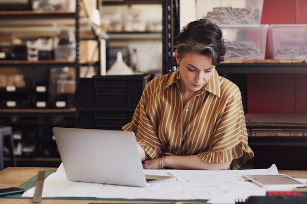 A woman business owner is seated at a desk with a laptop open in front of her. She has several paper spread out in front of her. She is inputting employee timesheets into her accounting software.