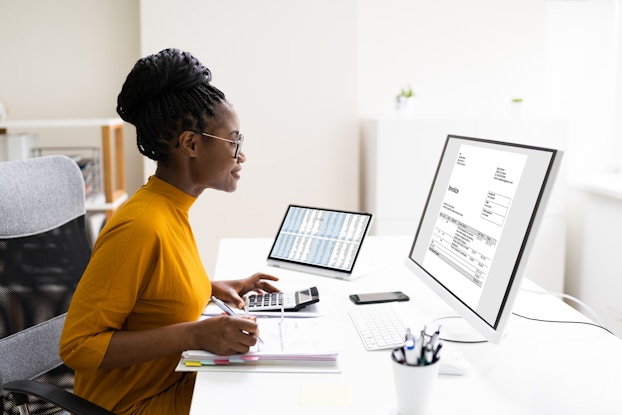 A woman sits at a large white desk and looks at a flatscreen computer monitor that shows an invoice. Her hands rest on a calculator and a binder with pages separated by pastel-colored tabs. To the woman's left is a tablet showing a table filled with rows of numbers.