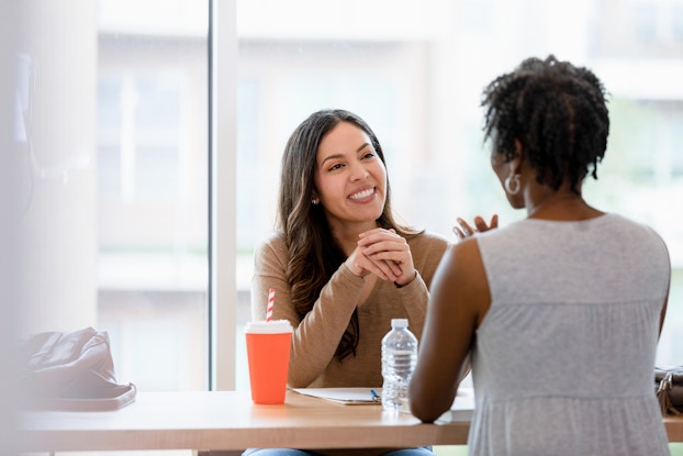 Two women sit across from one another at a table. The woman facing the viewer has long brown hair and is wearing a brown long-sleeved shirt. She has her elbows on the table and her hands folded, and she is smiling at the other woman. The woman facing away from the viewer has shirt dark hair and is wearing a light gray short-sleeved shirt and gold hoop earrings. Her expression cannot be seen, but she is gesturing with her hands.