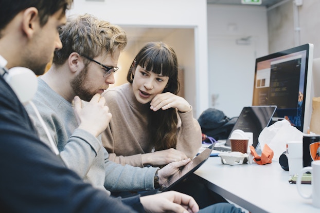  A profile short of three people sitting in a row at a desk, examining the electronic tablet in the hands of the man sitting in the middle. The man in the middle has blond hair and a dark beard. He has one hand held to his chin in thought. The person on the right, a woman with long, dark hair, is turned in the blond man's direction and is speaking. The person on the left, a dark-haired man, is in the foreground and slightly out-of-focus. On the desk in front of the three people are a computer monitor, an open laptop, and some trash from a food order.